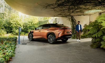 An orange Lexus RZ is parked and charging in a leafy green, vine-filled driveway. A man is seen walking around the rear of the car.