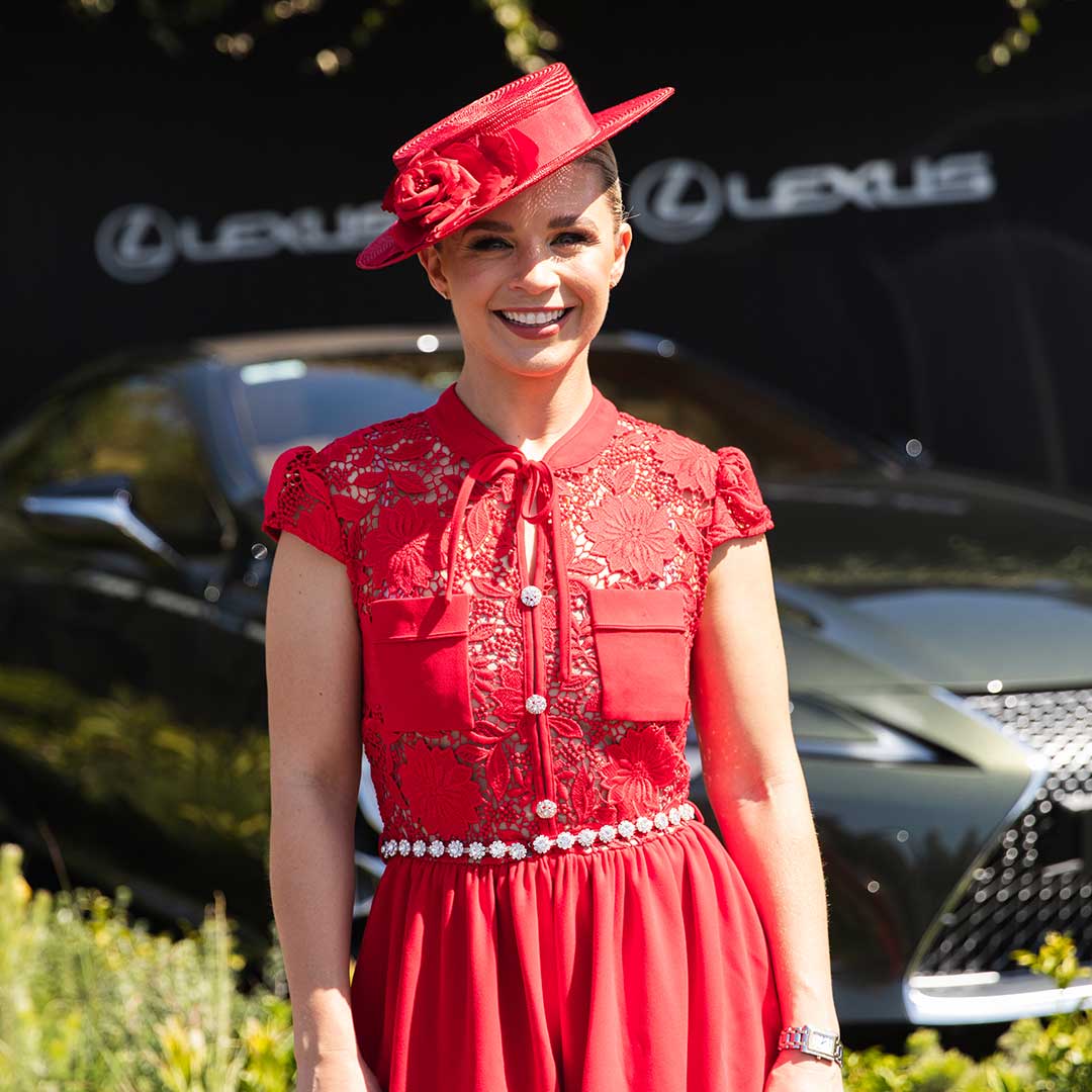 Racing presenter Emma Freedman smiles in a red lace dress and matching fascinator beside a Lexus at a race day event.