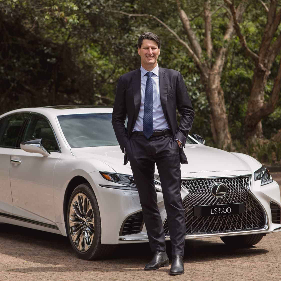 John Eales stands beside a white Lexus LS 500, wearing a dark suit and tie, outdoors with trees in the background