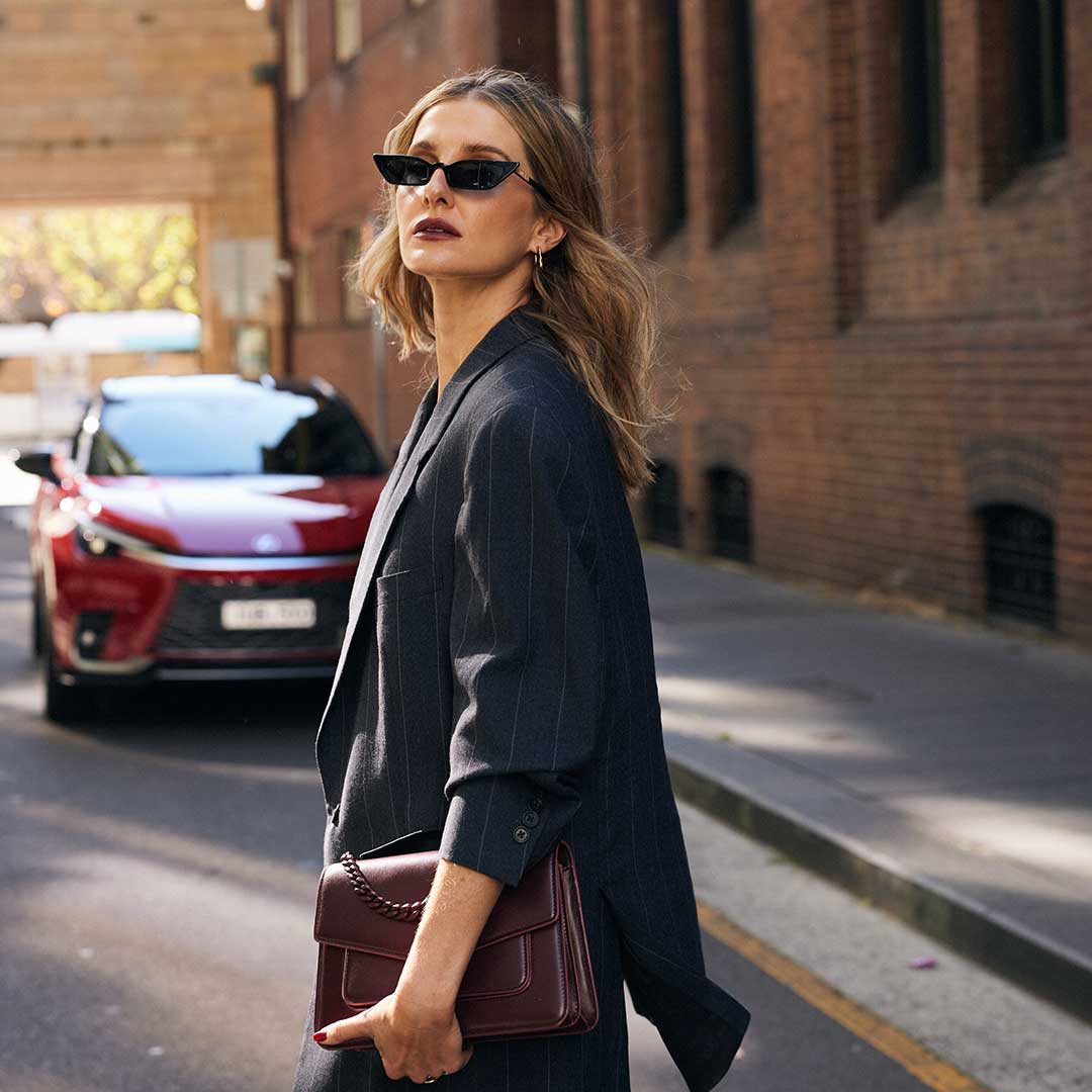 Kate Waterhouse poses in front of a red Lexus vehicle in a dark blazer and sunglasses on a city street.