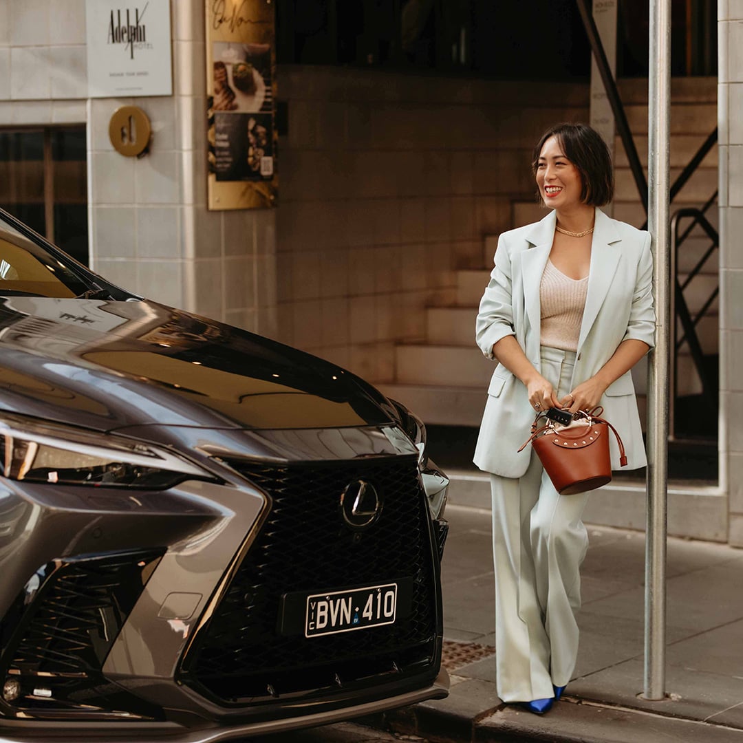 Melissa Leong stands  beside a Lexus vehicle on a city street in a pale blue suit, carrying a brown handbag.