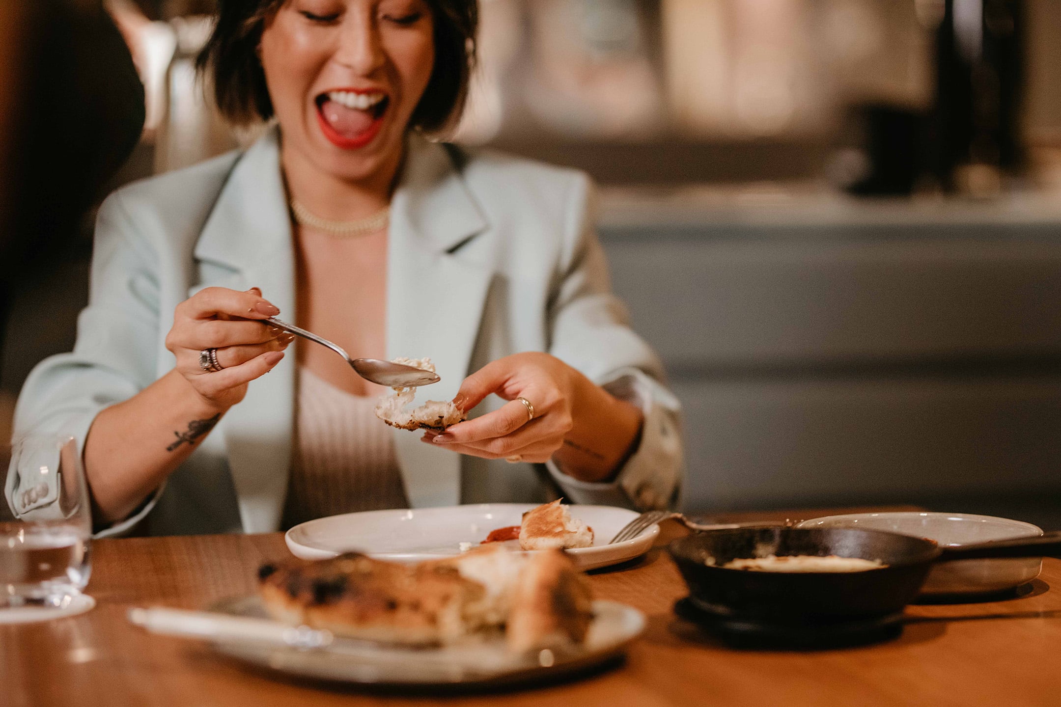 Melissa Leong laughs while enjoying a meal at a restaurant, wearing a pale blue blazer and seated at a table with food and drink.