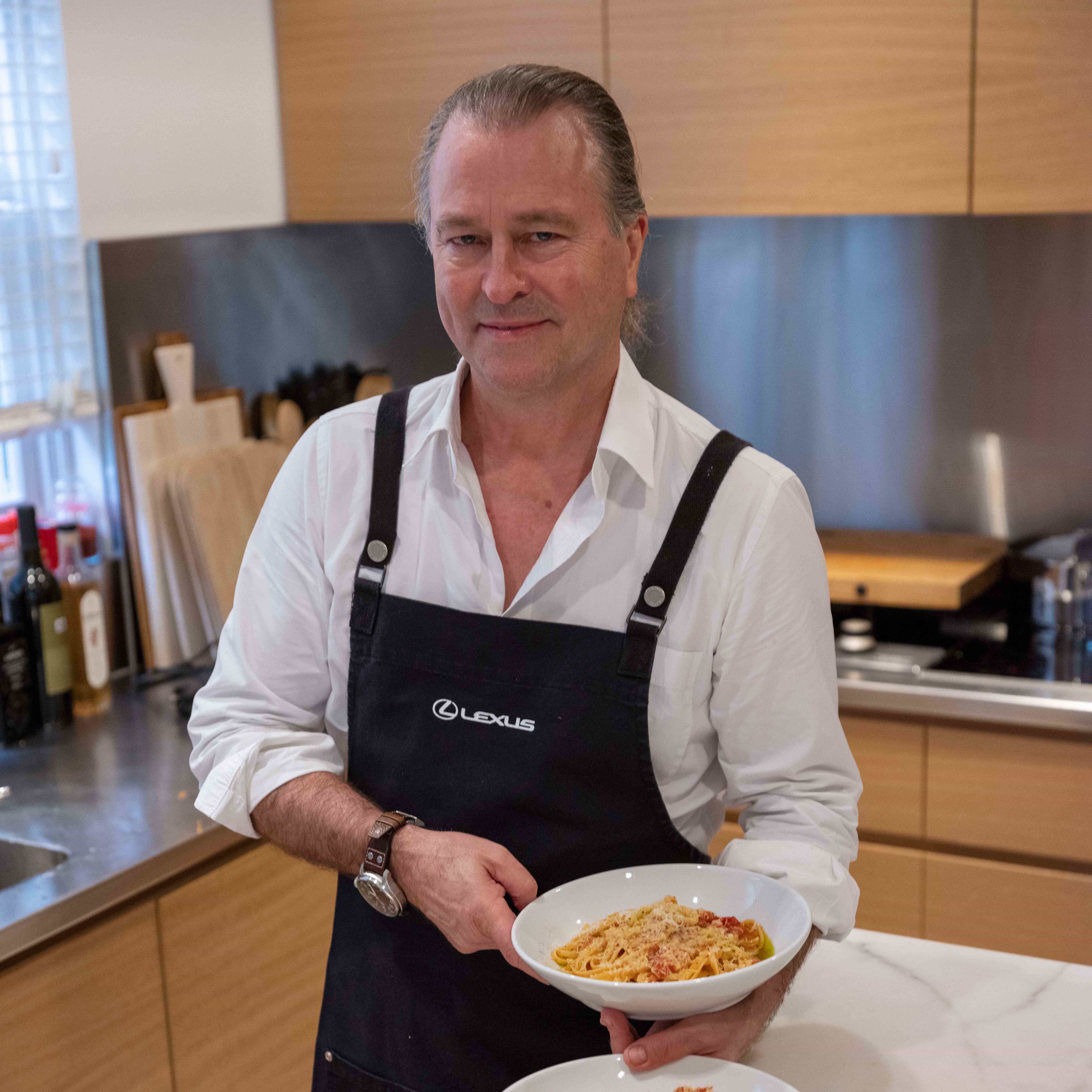 Neil Perry stands in a modern kitchen wearing a Lexus-branded apron, holding a bowl of pasta and smiling.