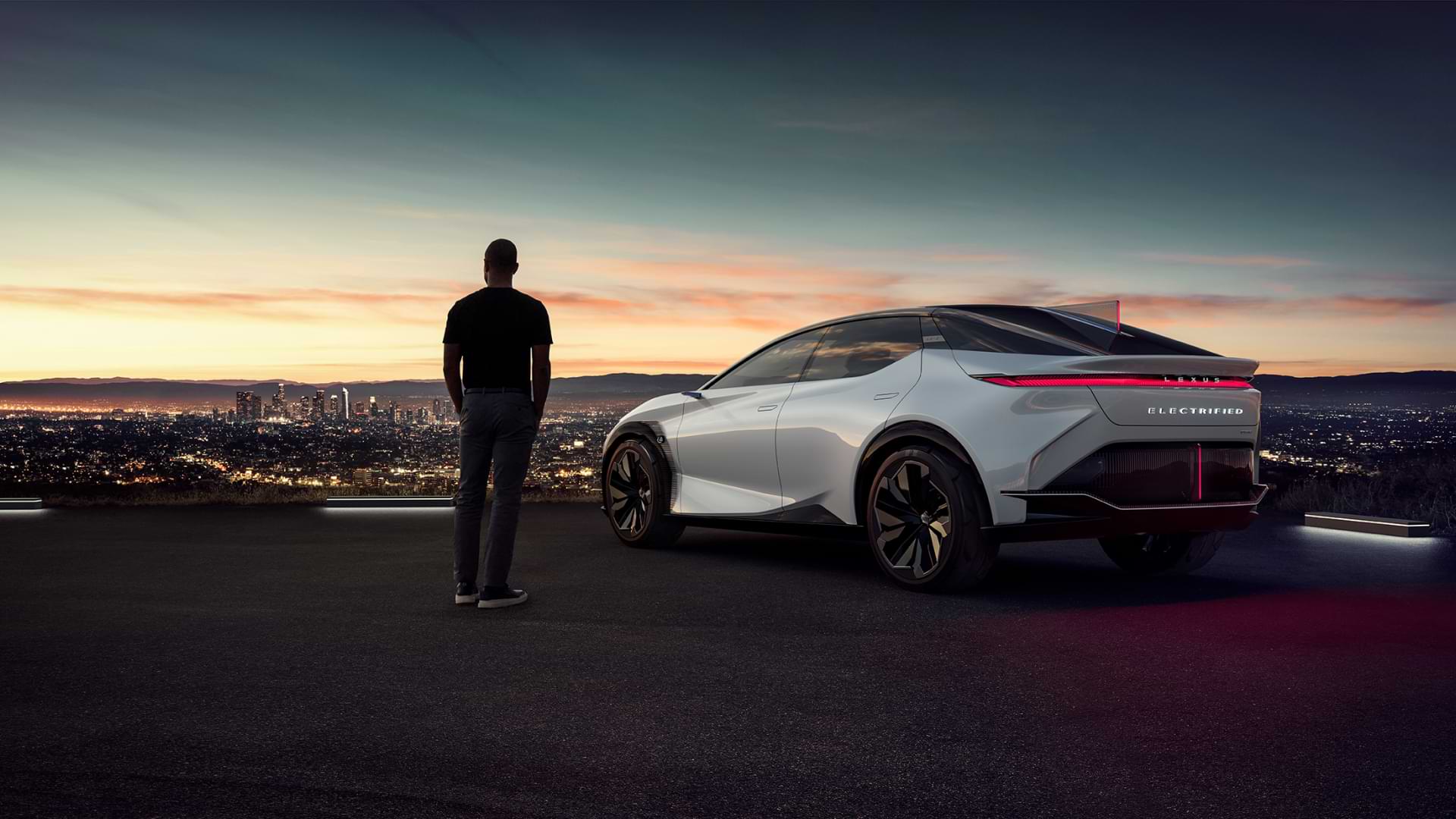A man standing next to the Lexus LF-Z Electrified concept vehicle at sunset overlooking a cityscape