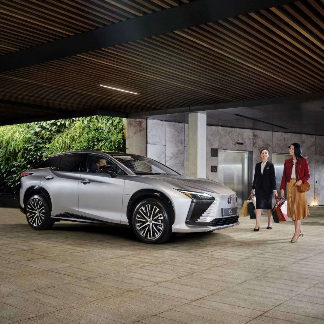 A silver Lexus RZ parked at a valet entrance as two women walking carrying shopping bags.