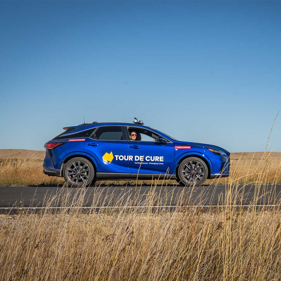 A blue Tour de Cure support vehicle drives along a rural road beneath a clear blue sky.