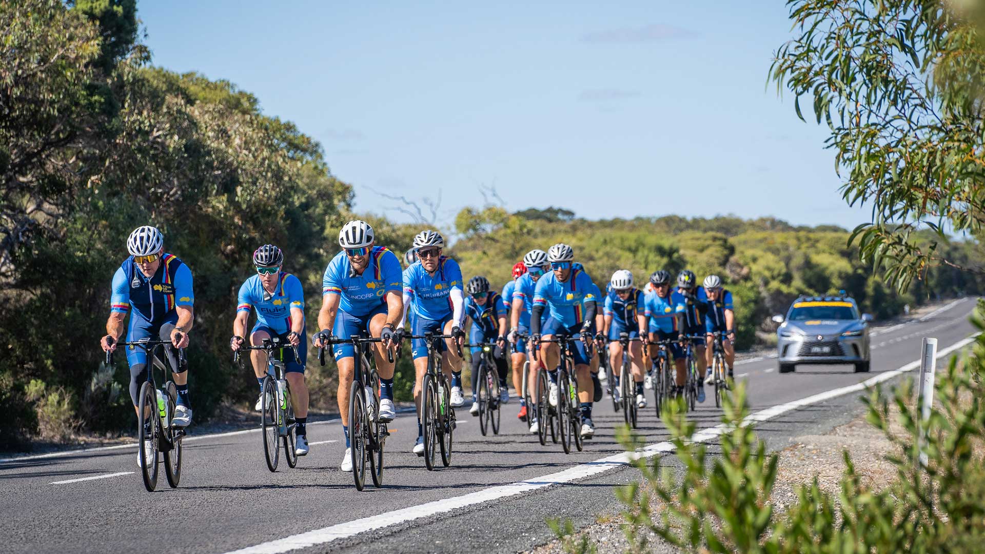 A group of cyclists ride together along a rural road, with a Lexus support vehicle following behind.