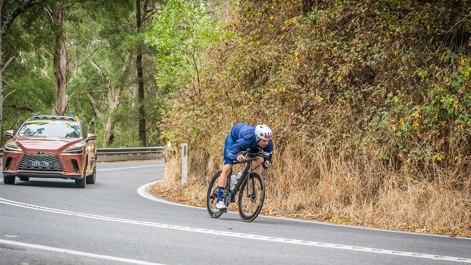 A cyclist rides uphill on a rural road with a Lexus support vehicle following behind.