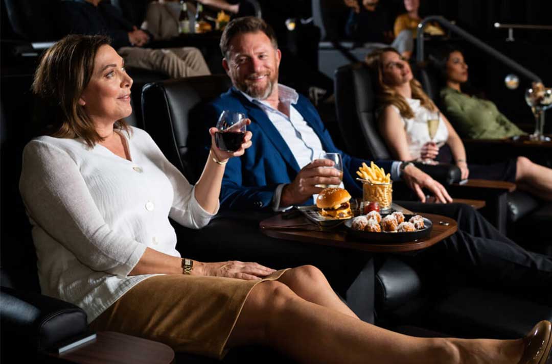 Guests sit in Gold Class cinema seats watching a movie, with food and drinks on side tables.