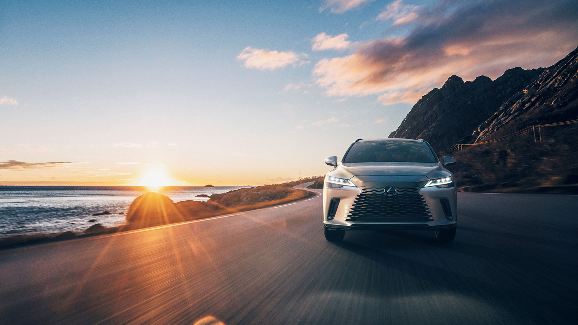 Front view of a Lexus SUV driving along a coastal road at sunset, with the ocean and cliffs in the background.
