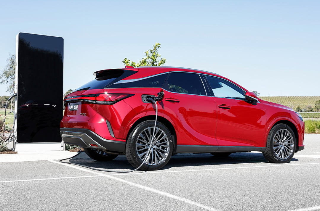 Red Lexus RX 450h+ plug-in hybrid charging at an electric vehicle station in an outdoor setting.