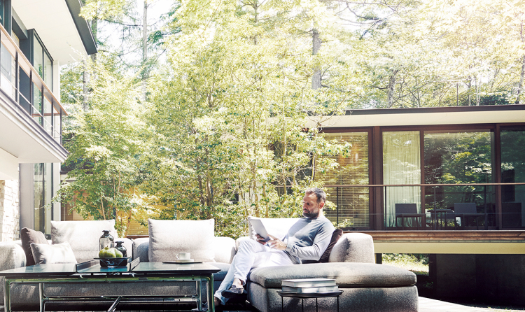 Man sitting on a couch in his backyard, reading a paper. Greenery surrounds him.