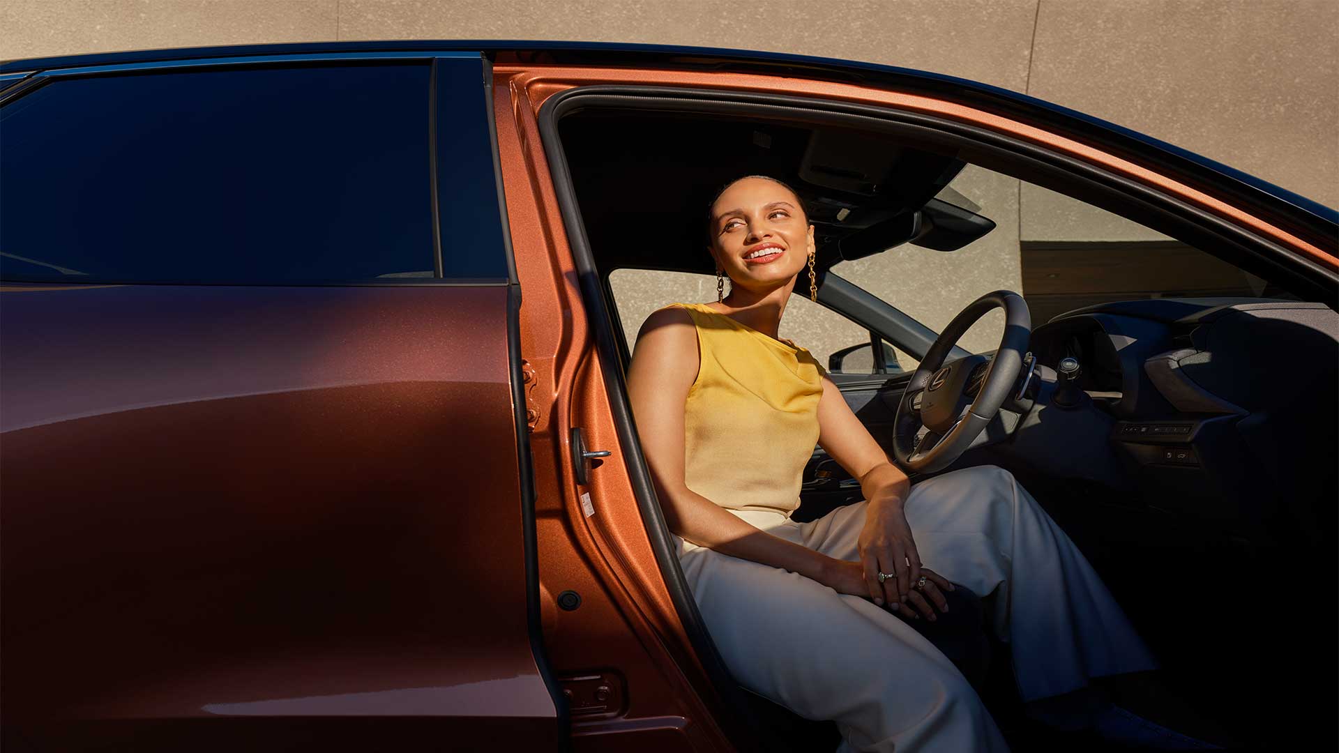 Woman sitting in the driver’s seat of a copper Lexus vehicle with the door open, smiling in sunlight.