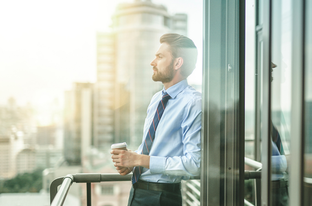 Corporate man stands on an office balcony with a coffee cup in his hand.