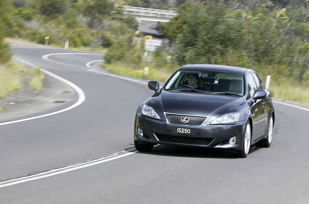 Black Lexus IS 250 driving along a winding road with trees and flowers in the background