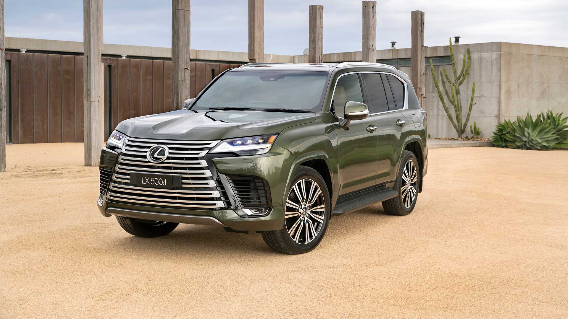 A green Lexus LX parked on a sandy driveway with wooden posts in the background and a cactus