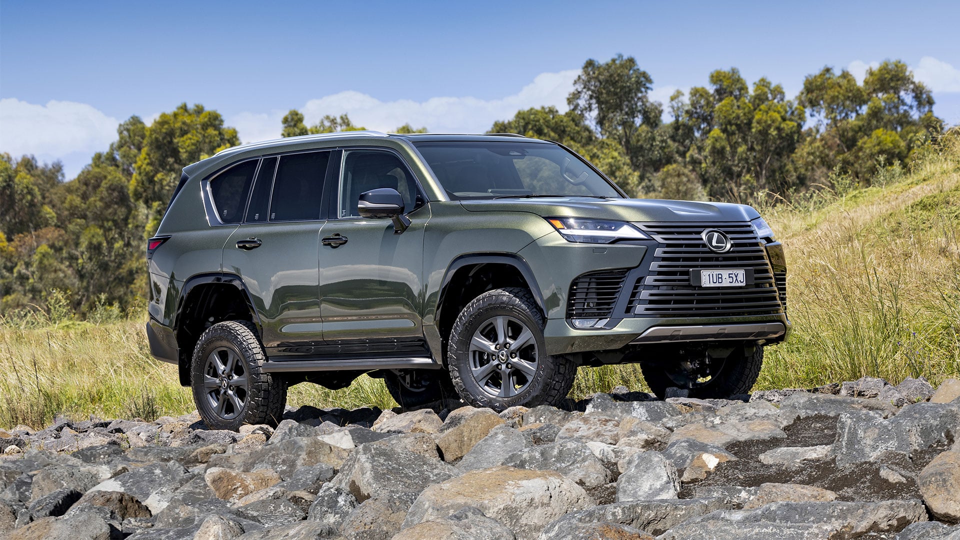 A green Lexus LX Overtrail parked on top of rocks with trees and a blue sky in the background