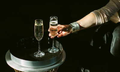 Woman with silver bracelet reaches for a glass of champagne near another full glass on a dark table.